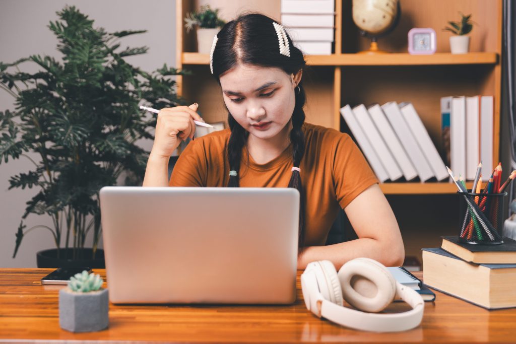 A woman with braided hair studies intently on a laptop at a desk surrounded by books, colored pencils, and headphones