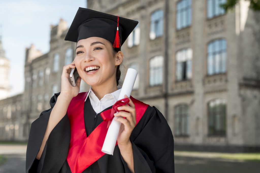 A young woman in graduation cap and gown smiles while talking on the phone, holding a rolled diploma outside a university building