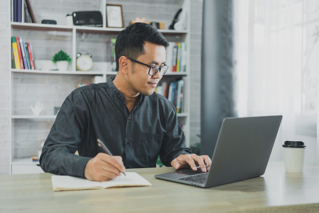 A man in glasses takes notes while working on a laptop at a wooden desk with a bookshelf in the background