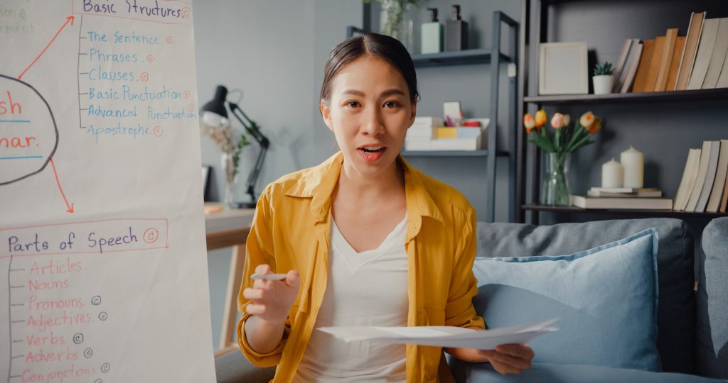 A young woman in a yellow shirt teaches English grammar online from her home, referencing a flipchart with notes on basic structures and parts of speech