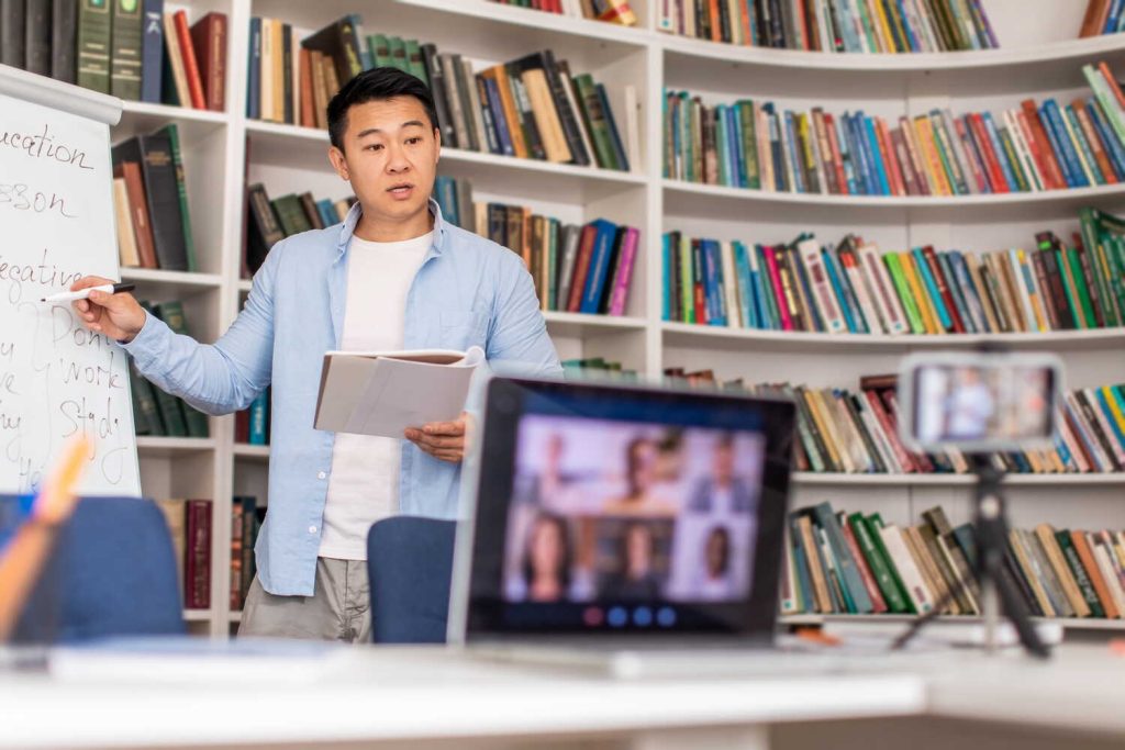 A male teacher conducts an online class from a library, writing on a whiteboard while students join via video call