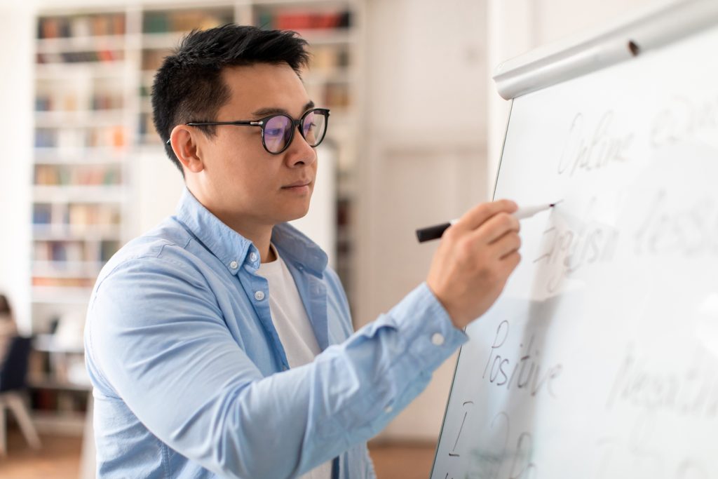 A professor wearing glasses writes grammar rules on a whiteboard in a classroom setting