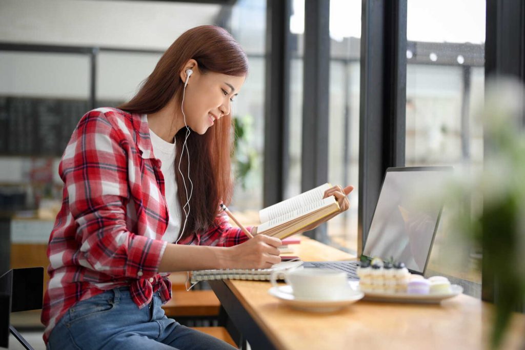 A woman in a plaid shirt taking notes from a textbook while listening through earphones, with a laptop open on a café table beside her
